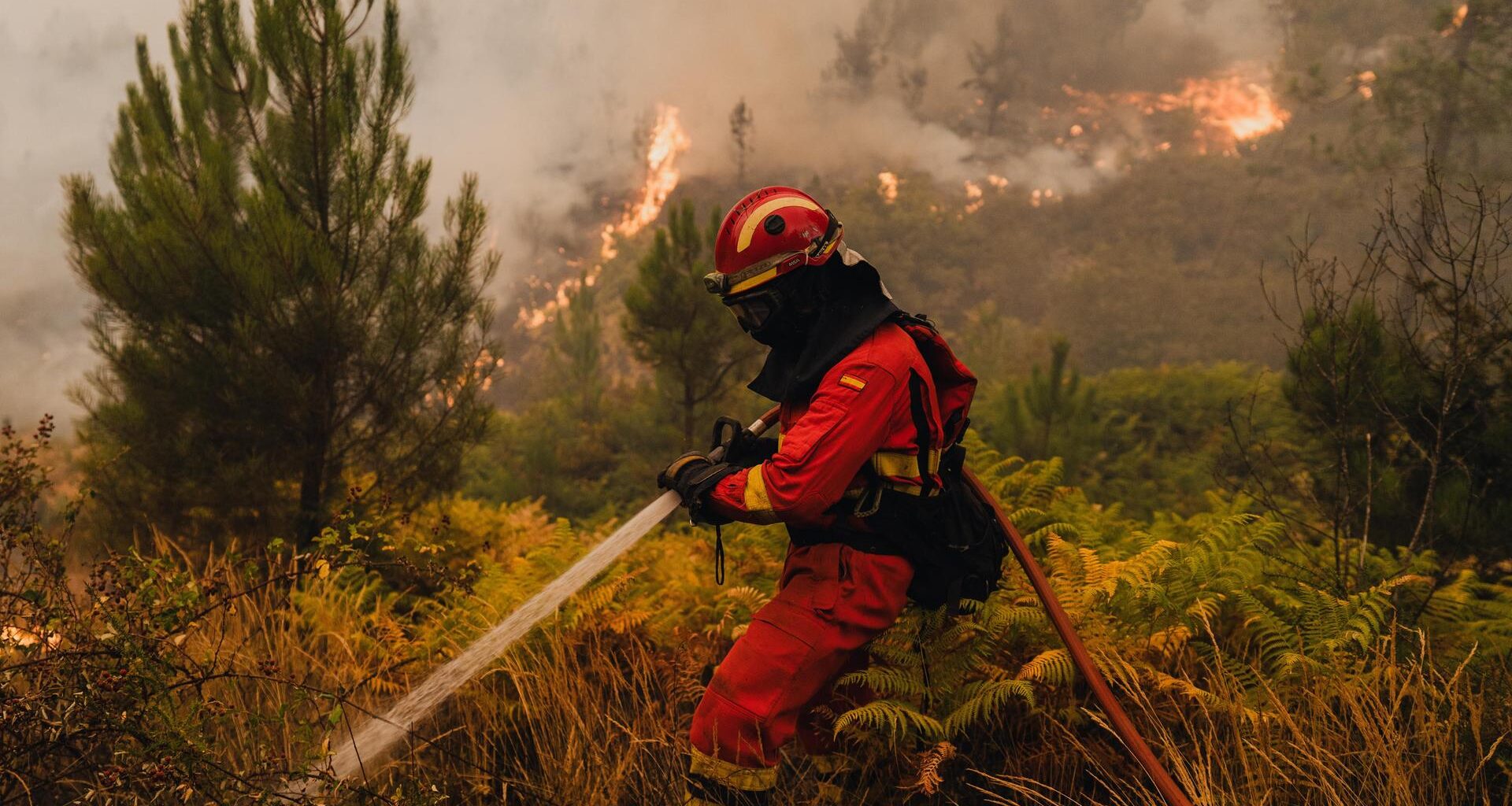 Ein Mann in Feuerwehr-Uniform hält einen Wasserschlauch und versucht eine brennende Fläche zu löschen.