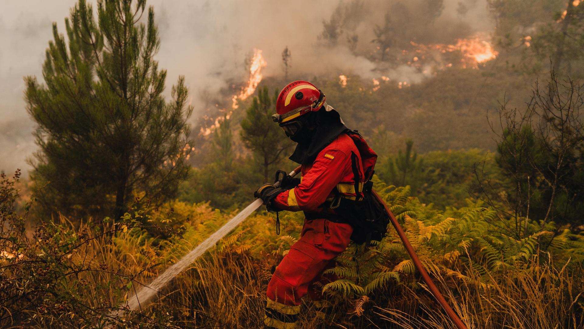 Ein Mann in Feuerwehr-Uniform hält einen Wasserschlauch und versucht eine brennende Fläche zu löschen. Ein Mann in Feuerwehr-Uniform hält einen Wasserschlauch und versucht eine brennende Fläche zu löschen.