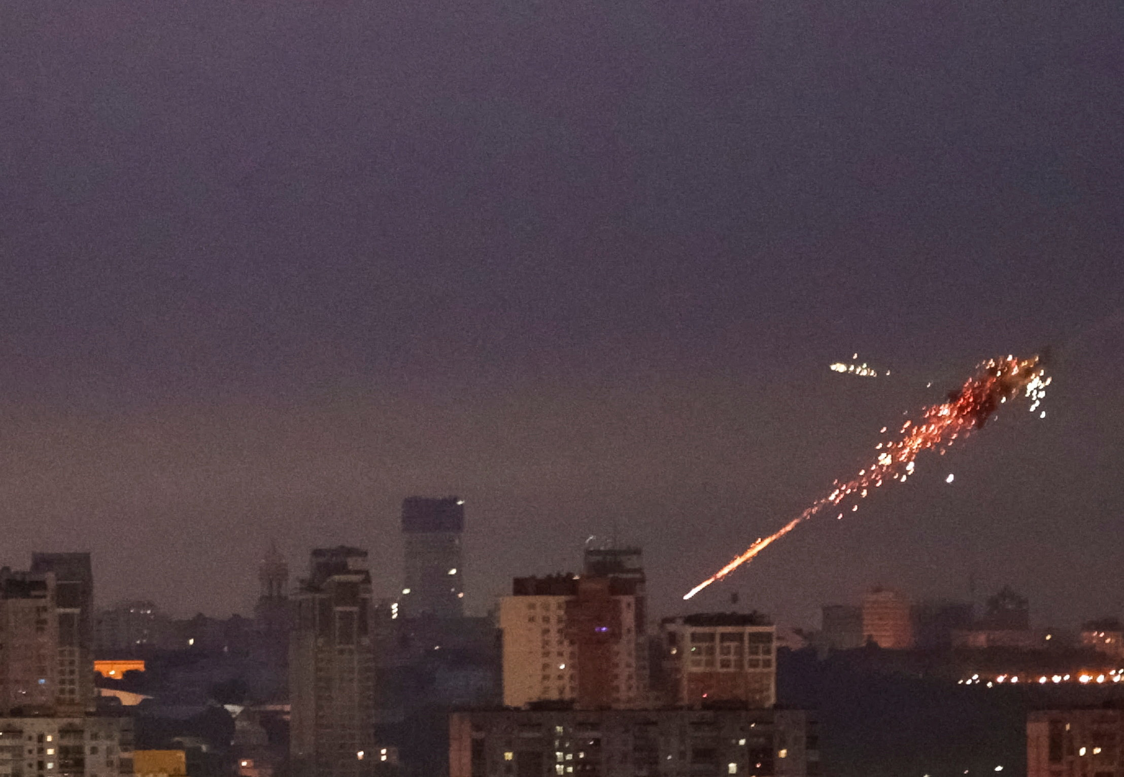 A missile exploding in the night sky over Kyiv, with sparks trailing from the explosion and city buildings visible below.