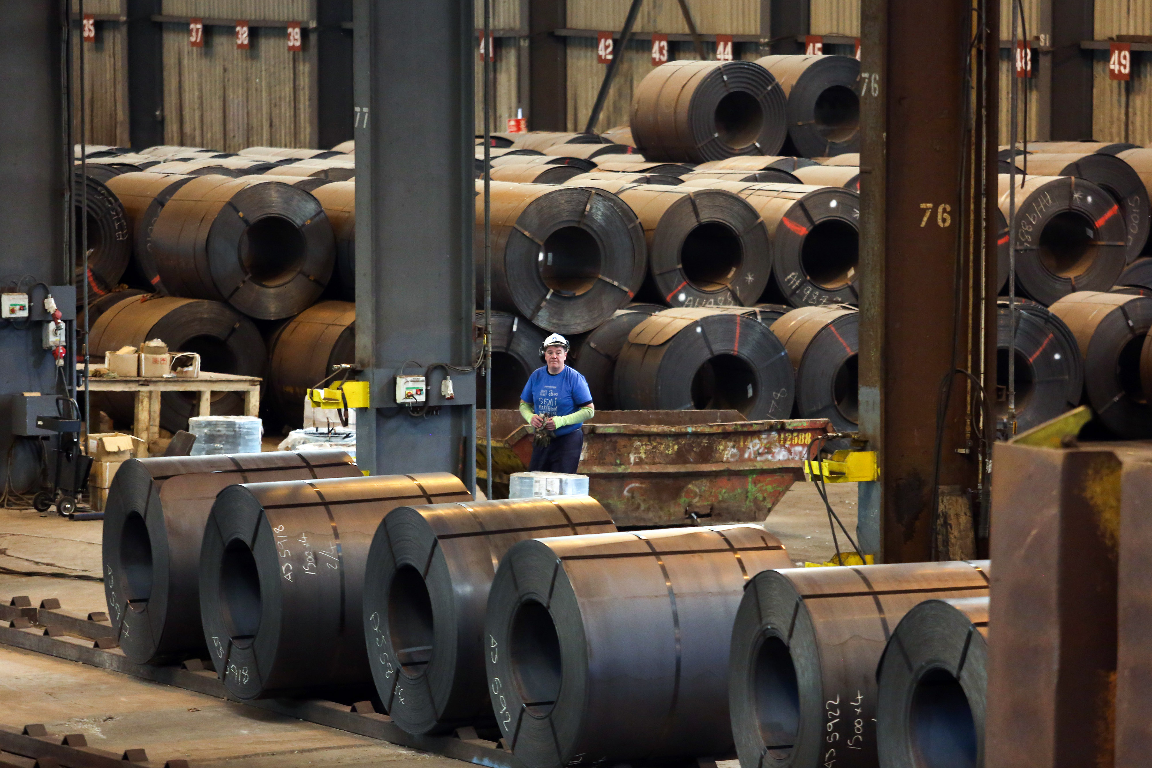 An employee standing in a steel mill with large coiled sheets of metal.