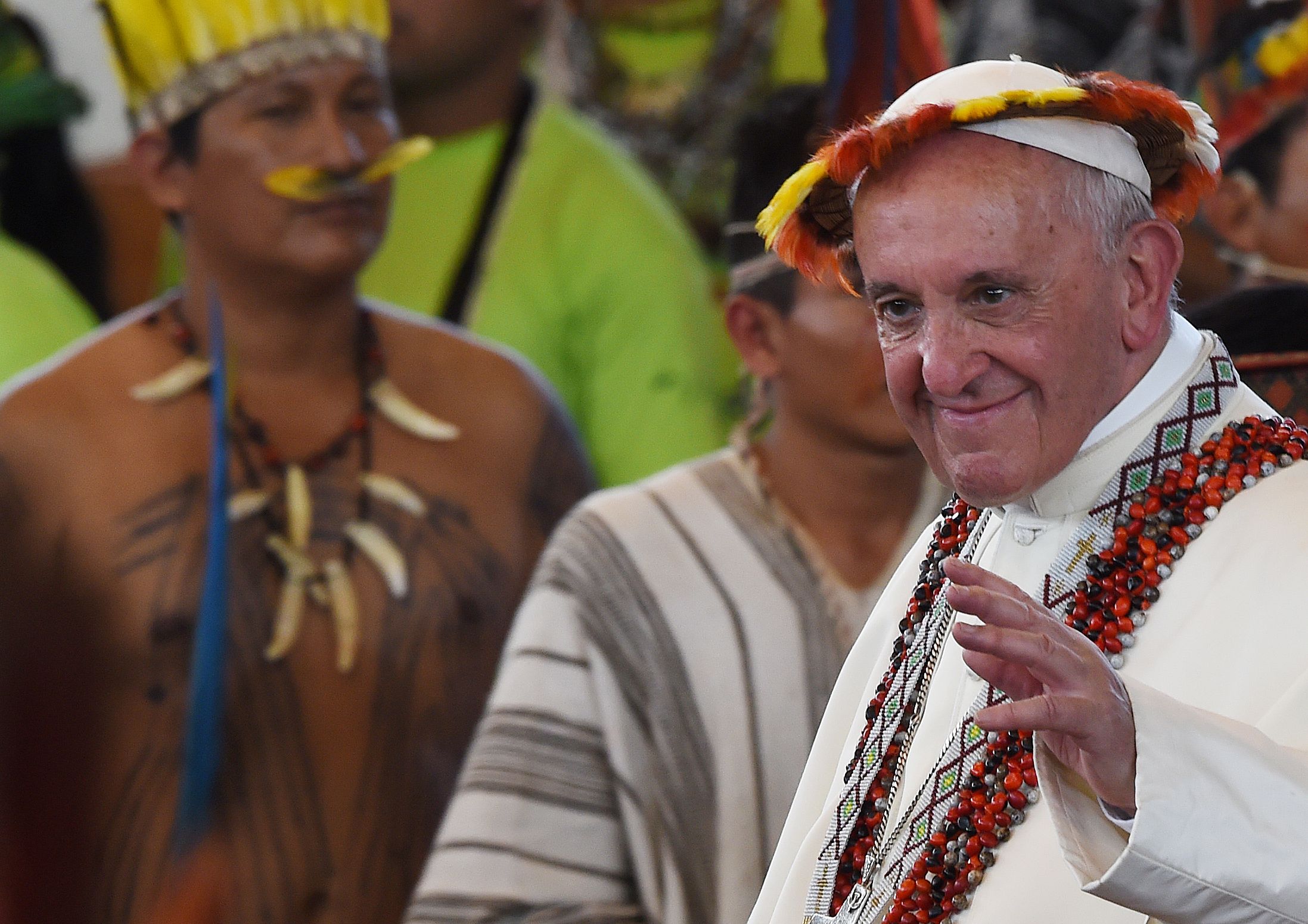 Pope Francis wearing indigenous headdress and necklace, waving.
