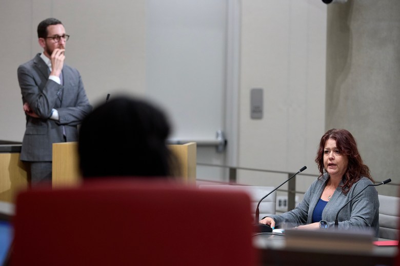 A person sits at a microphone testifying or speaking during a public meeting or hearing, while another person in a suit stands in the background with a thoughtful expression. The scene takes place in a formal setting with modern furniture and a concrete wall.