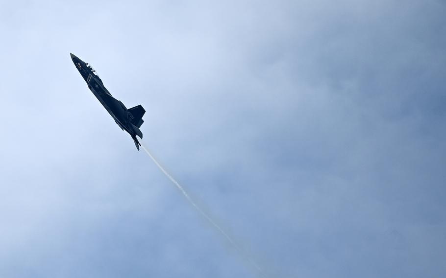 A dark military fighter jet performing a steep climb or aerobatic maneuver against a cloudy blue sky, with a visible vapor trail streaming behind the aircraft.”