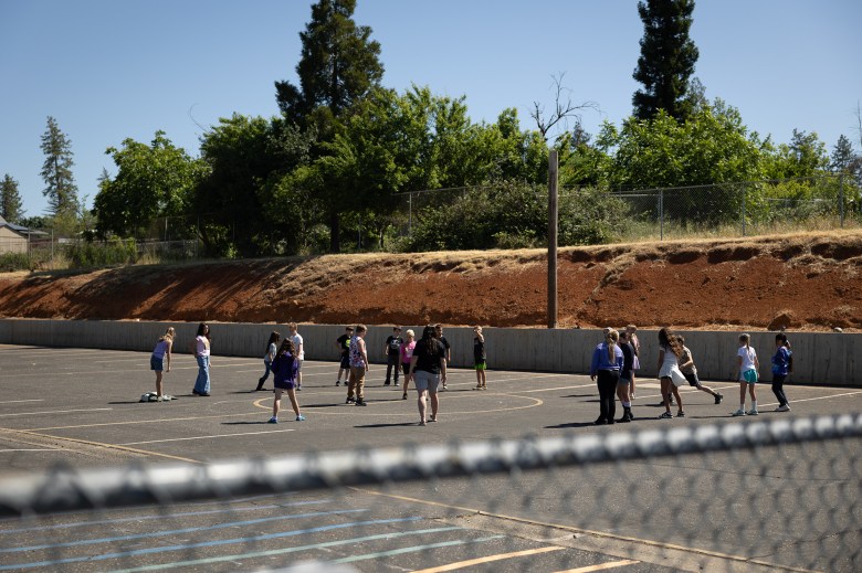 A wide view of students playing on an asphalt playground on a clear day. Trees, dirt and some growing vegetation can be seen nearby. The top of a wired fence can be seen in the foreground.