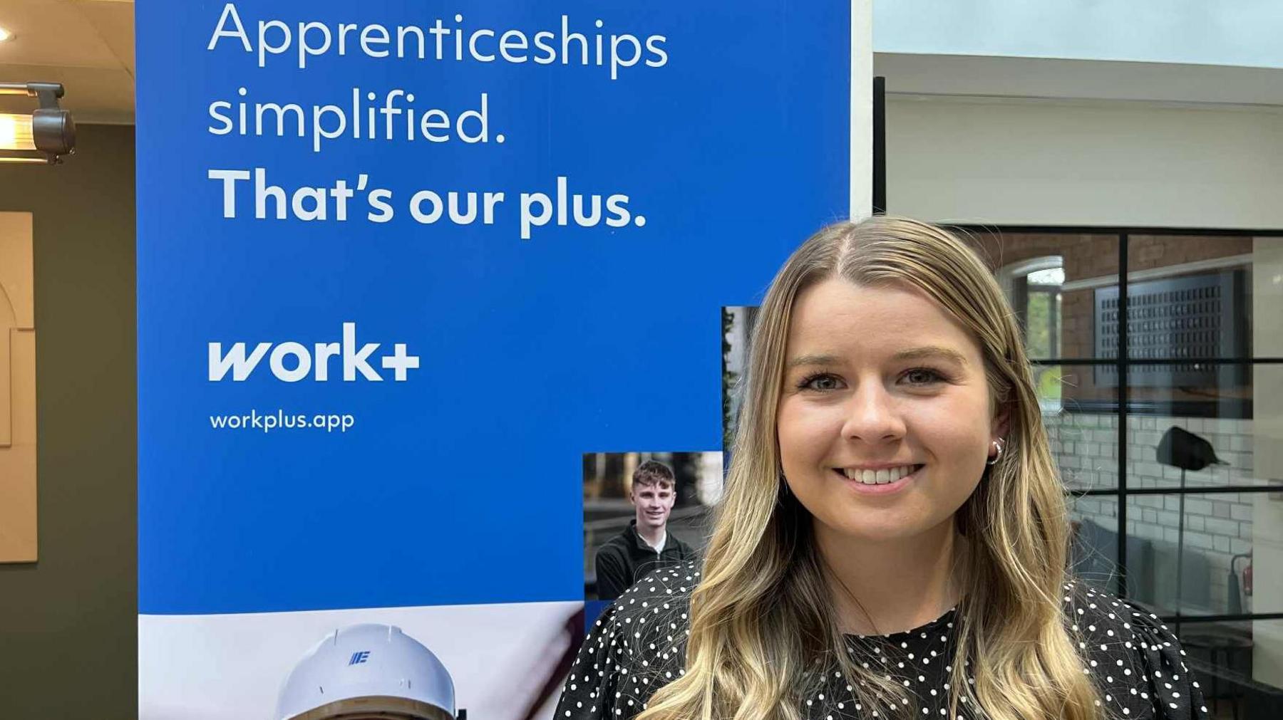 A woman with blonde hair is wearing a polka dot top with a large promotional board behind her left shoulder says "Apprenticeships simplified. That's our Plus.
Work+
Workplus.app"

On her right shoulder a large window is in the background