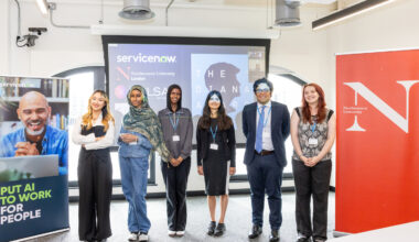 Six graduates of Northeastern University’s fast-track digital course pose side by side in a classroom, smiling in front of a projector screen and banners for ServiceNow and Northeastern. The cohort includes men and women in professional attire, representing a diverse group.