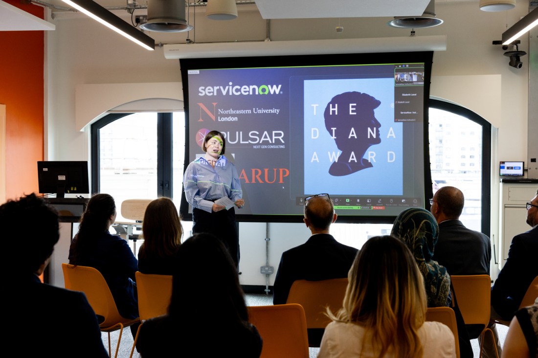 A woman stands at the front of a classroom at Northeastern University London, speaking to an audience seated in rows of orange chairs. Behind her, a large presentation screen displays logos of partnering organizations including ServiceNow, The Diana Award, Pulsar, and Arup. The screen also shows a Zoom call interface with the Diana Award logo featuring a silhouette of Princess Diana. The audience includes a diverse group of individuals, attentively listening to the presentation. Bright daylight filters in through large arched windows.