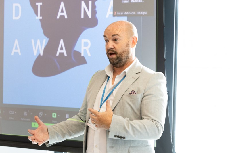A speaker in a light gray blazer gestures while presenting at a graduation event for Northeastern’s London campus. Behind him is a large screen with The Diana Award logo.