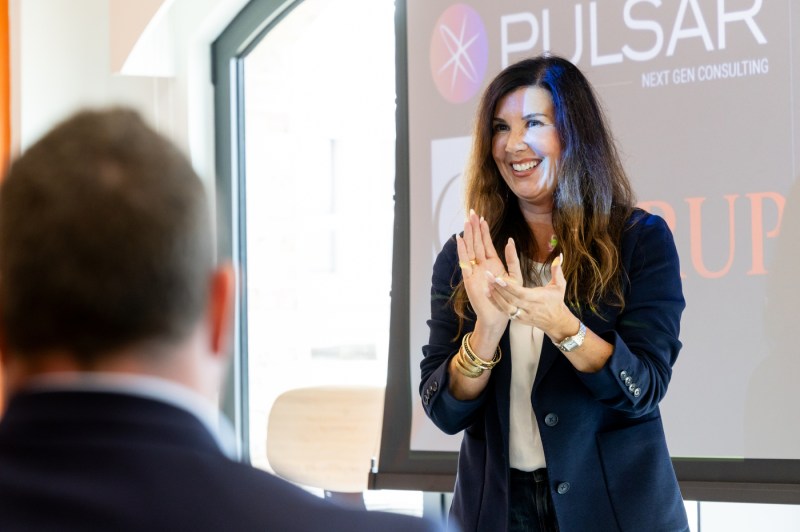 A woman claps and smiles while standing in front of a screen showing the logos for Pulsar, The Diana Award, and ServiceNow during a graduation ceremony at Northeastern University’s London campus. A man stands beside her as audience members watch.