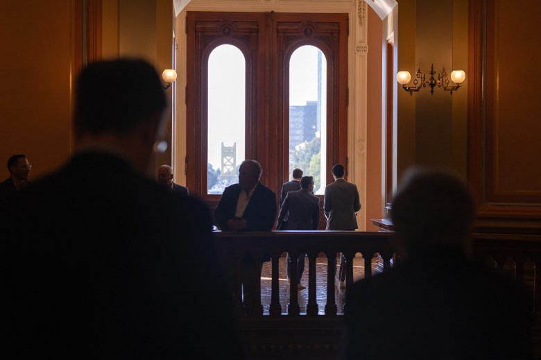 Two giant doors with oval glass windows illuminate the silhouettes of people standing around and talking to each other at the state Capitol in Sacramento.