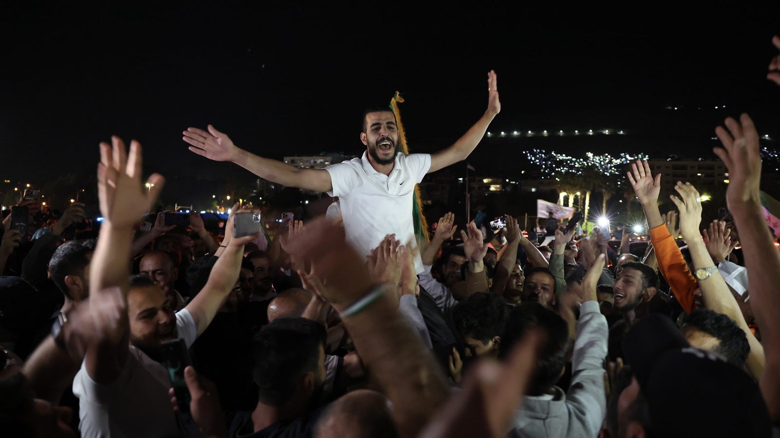 A crowd of people joyfully celebrates at night, with one individual in a white shirt being held aloft by others. Many attendees have their hands raised, and some are recording the event with smartphones.