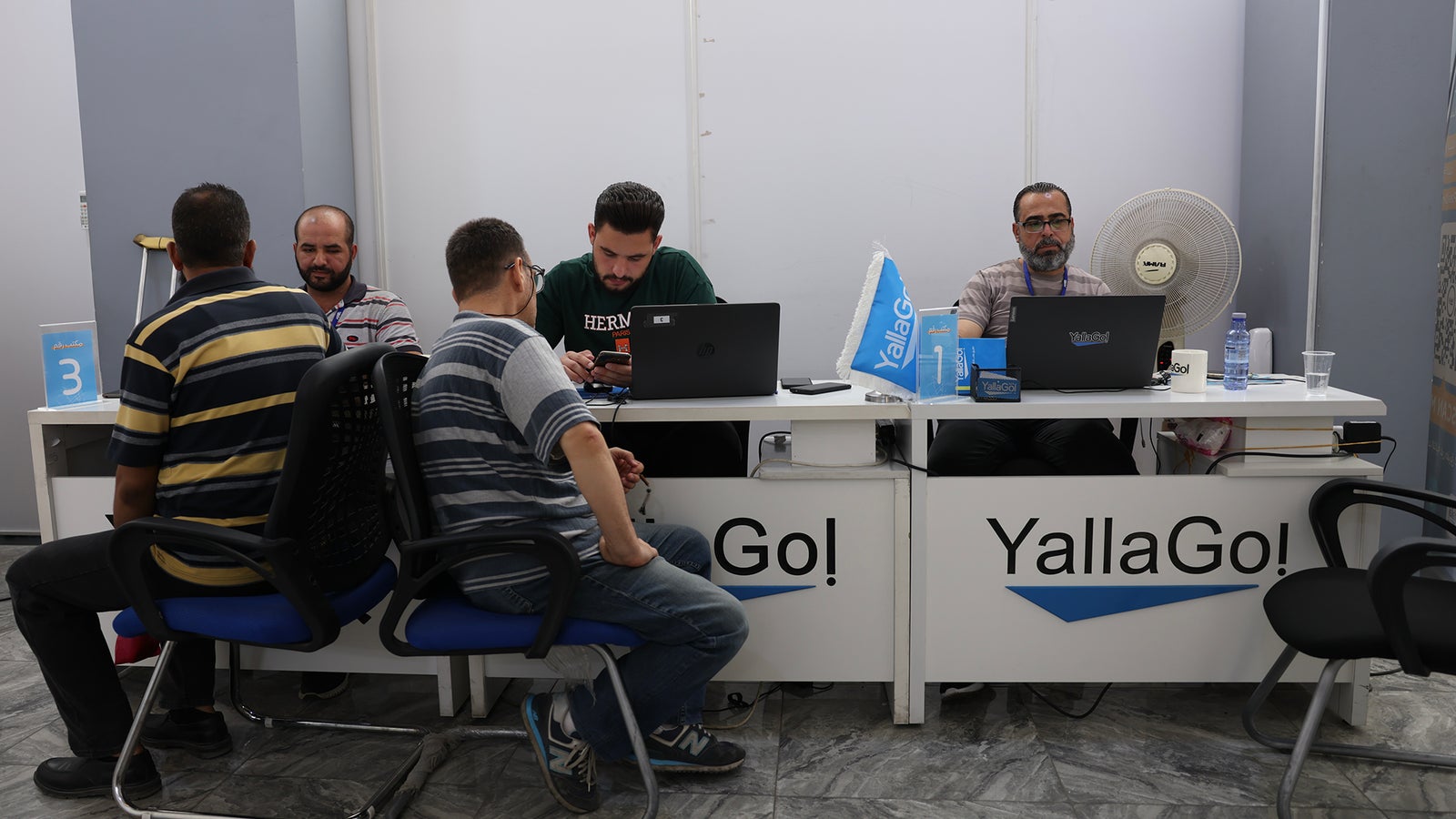 A service desk area with multiple individuals seated. Two men in striped shirts are conversing, while three others are engaged with laptops. The desk displays the logo "YallaGo!" and includes a fan and water bottles. A sign indicating the number "3" is visible in the background.