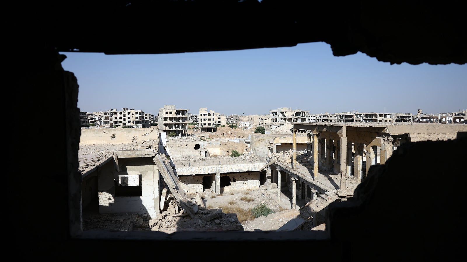 A view through a broken wall into a desolate landscape of ruined buildings, with collapsed structures and debris scattered across the ground under a clear blue sky.