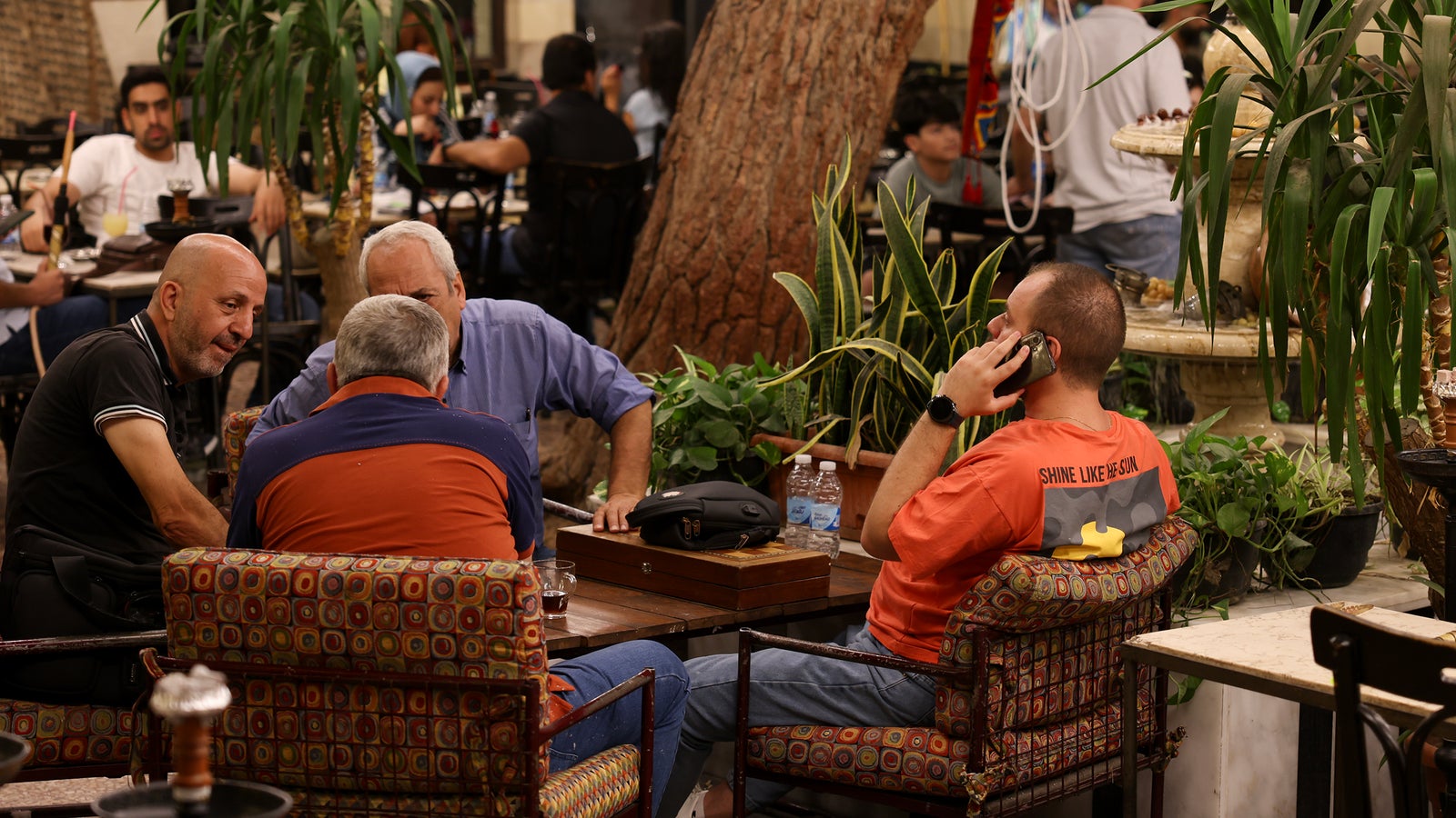 A group of four men are sitting around a colorful, patterned table in a lively café with indoor greenery. One man, wearing an orange shirt, is on a phone, while the others are engaged in conversation. In the background, a fountain can be seen and patrons are scattered throughout the café.