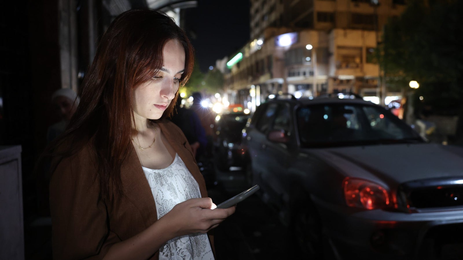 A young woman with long brown hair, dressed in a white top and brown jacket, is intently looking at her smartphone in an urban nighttime setting. The background features blurry street lights and parked cars, indicating a busy city scene.