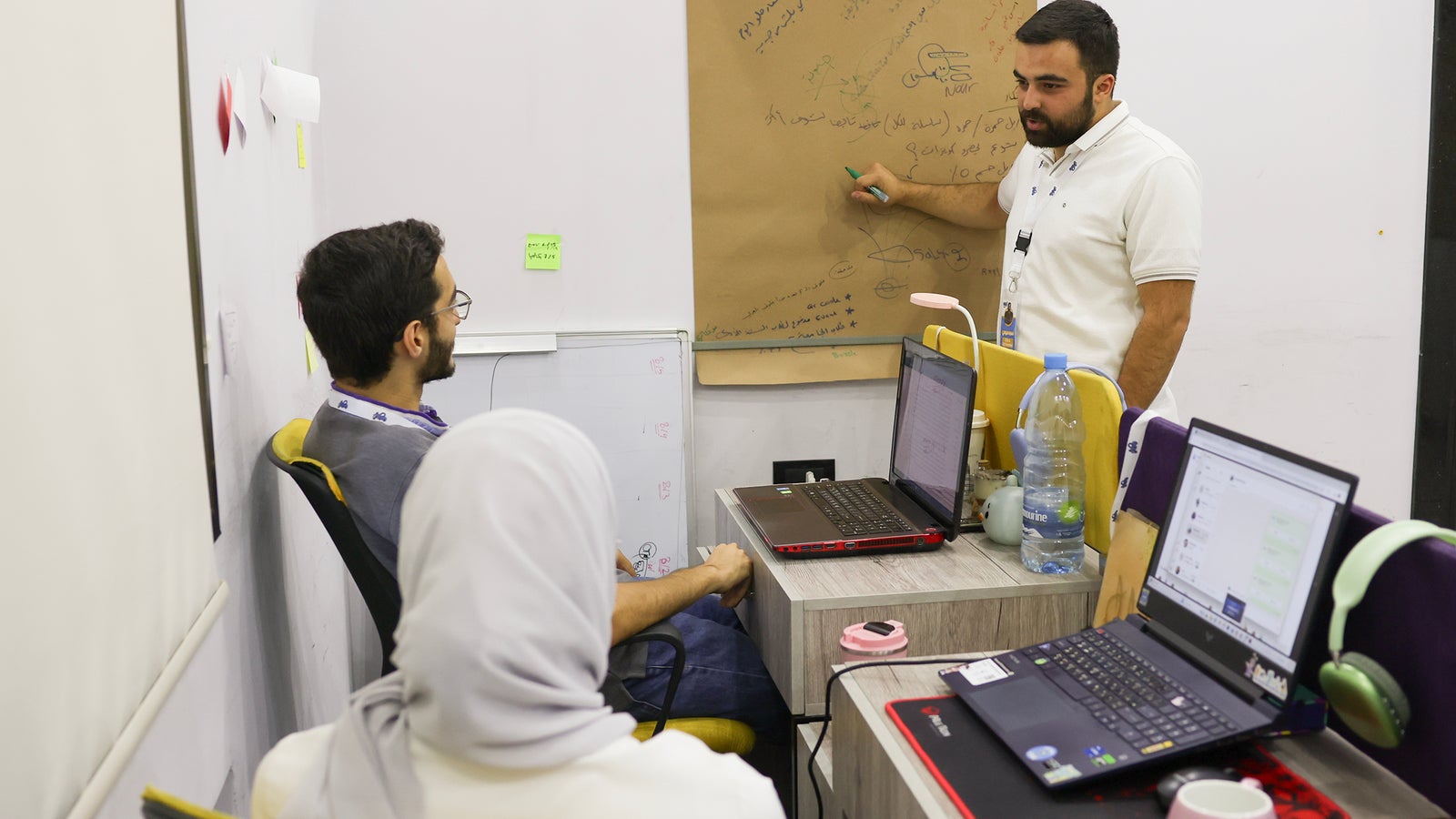 A group meeting in a modern office setting where a man in a white polo shirt is standing beside a wall covered with brown paper and notes, explaining a concept; two seated individuals, one wearing a hijab, are attentively listening with laptops open on their desks.