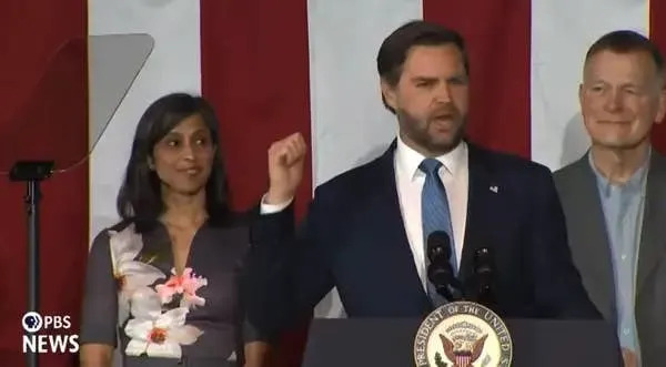 A man in a suit speaks at a podium with the U.S. seal, flanked by two other people, in an event setting