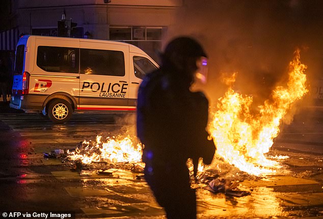 A police officer in riot gear walks past a fire in a street in Lausanne, on August 25, 2025