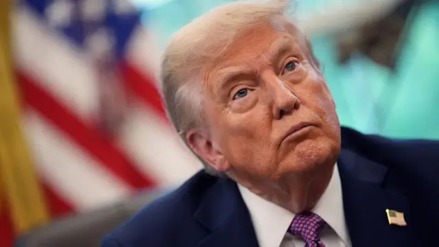 Imagen de cerca del presidente estadounidense, Donald Trump, quien lleva un traje azul oscuro, camisa blanca y corbata morada con lunares azules. También lleva una insignia estadounidense en la solapa derecha de su chaqueta.