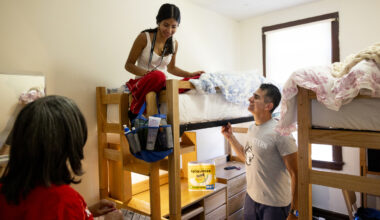 A student sits on top of a bunk bed unpacking while their family members stand in their dorm room with them.