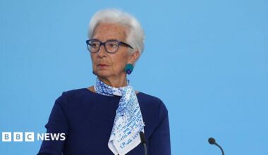 Christine Lagarde, president of the European Central Bank, standing at lectern on a stage. She is wearing a blue dress with a blue and white patterned scarf. she wears glasses, and has short, white hair