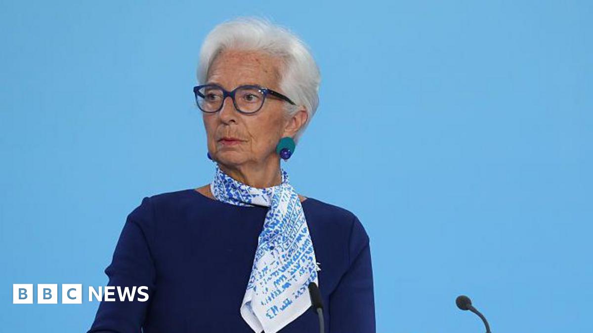 Christine Lagarde, president of the European Central Bank, standing at lectern on a stage. She is wearing a blue dress with a blue and white patterned scarf. she wears glasses, and has short, white hair