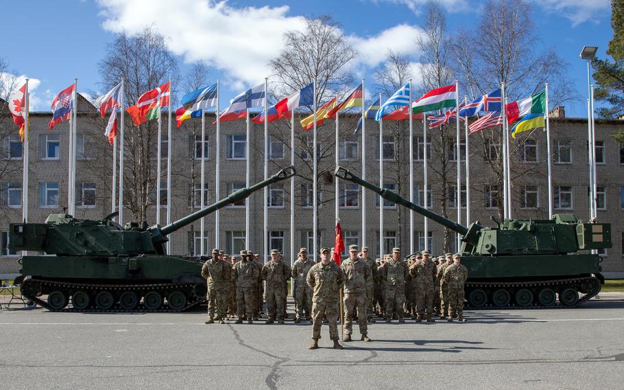 Soldiers stand at ease as national flags fly in the background.