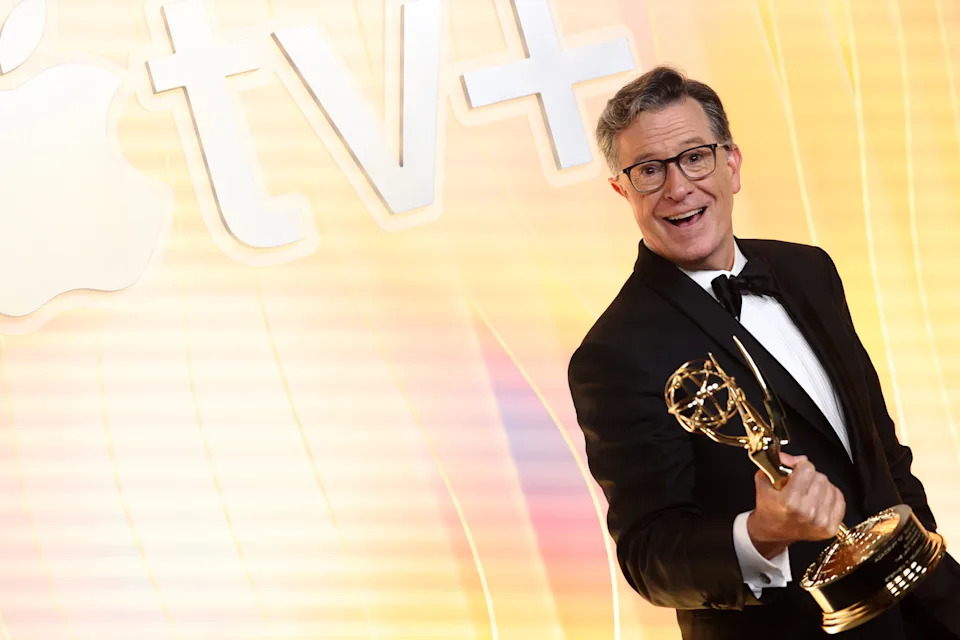 Person in tuxedo holding an award trophy, smiling on a stage with Apple TV+ branding in the background