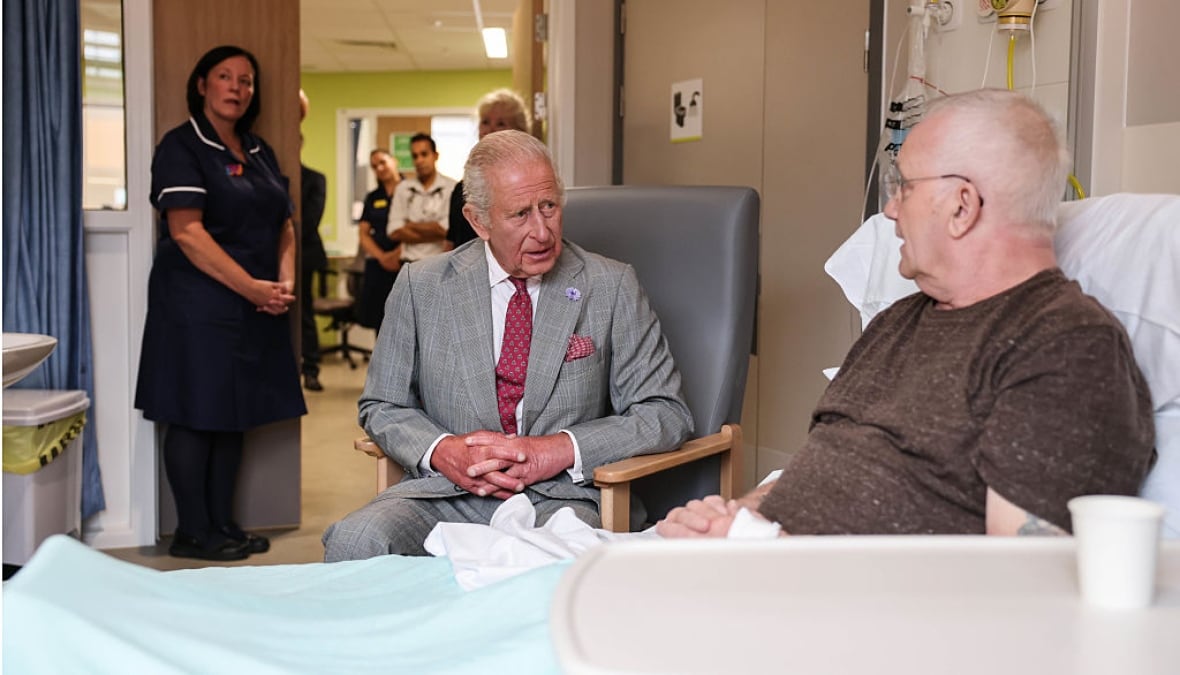 A person sits beside a person in a hospital bed and talks to them as another person looks on.