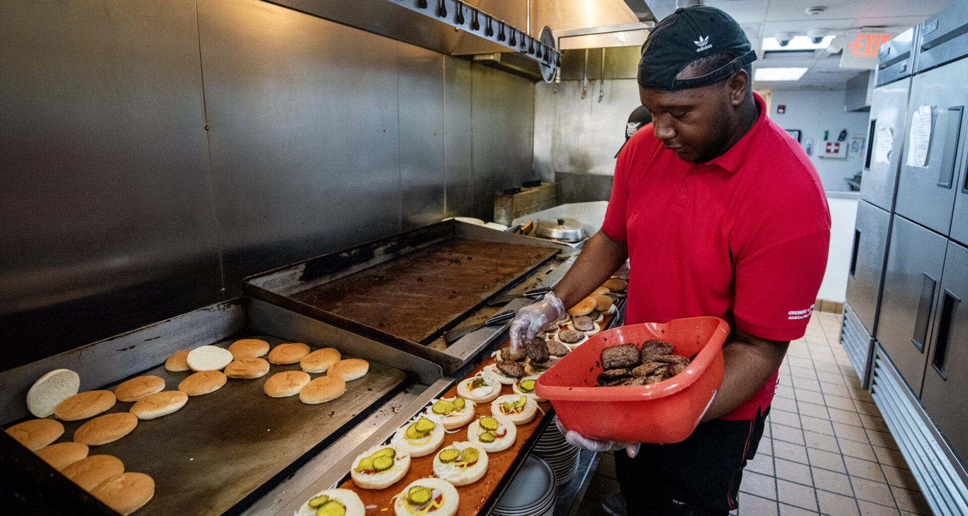A cook in a red shirt and cap prepares burgers in a commercial kitchen, placing cooked patties on buns lined up on a counter.