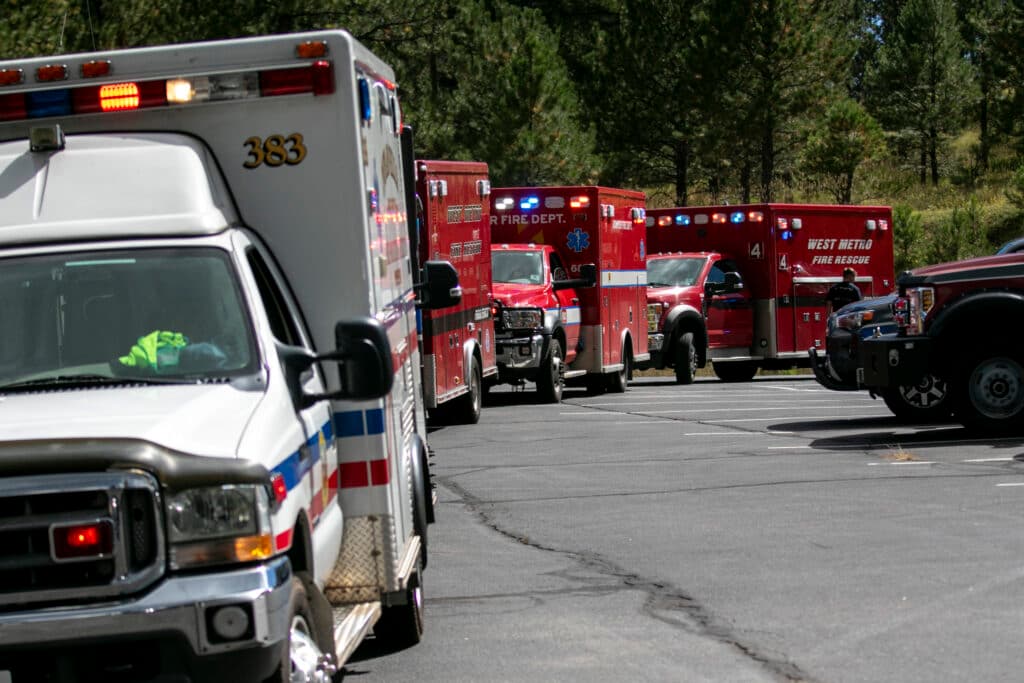 A row of emergency vehicles, including ambulances and fire trucks, is parked on a paved road surrounded by trees. Their emergency lights are flashing.