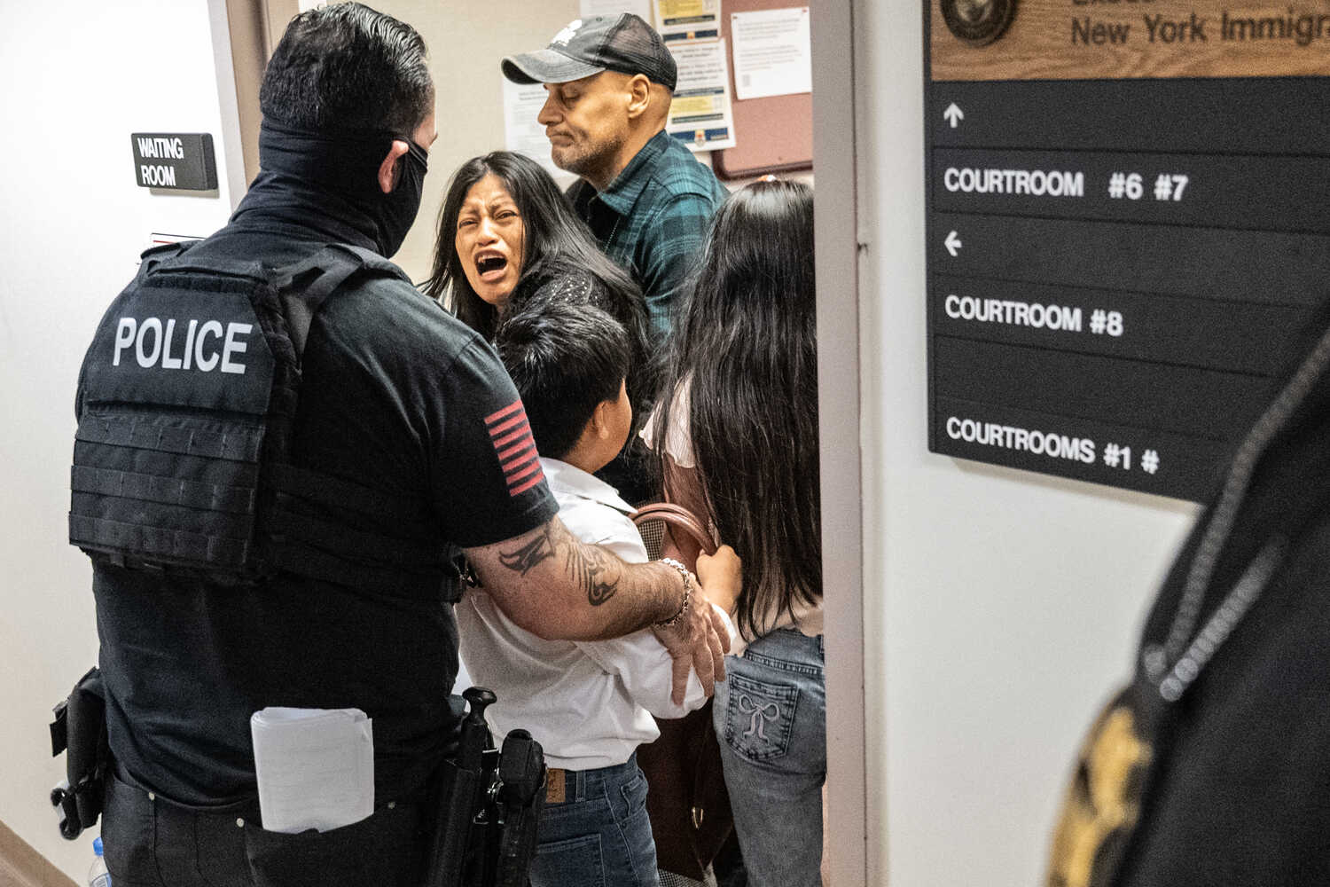 A woman is restrained by a man in a plaid shirt as a man wearing a police vest holds a little boy, and a young girl stands next to the boy.