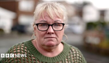 Mandy - a woman with blond hair, gold earrings and black-rimmed glasses. She is wearing a dark-green sweater. She is standing on a street.