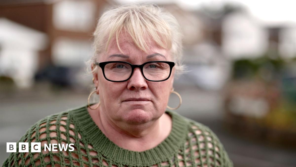 Mandy - a woman with blond hair, gold earrings and black-rimmed glasses. She is wearing a dark-green sweater. She is standing on a street.