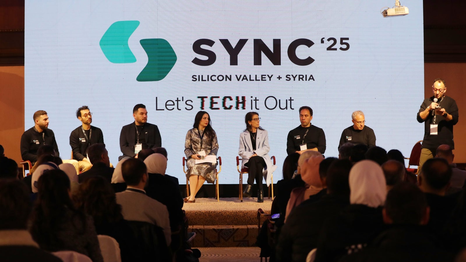 A panel discussion at the SYNC '25 event featuring seven speakers seated on stage. The backdrop displays the event logo and the phrase 'Let's TECH it Out'. Attendees are visible in the foreground, and some audience members are wearing light-colored head coverings.