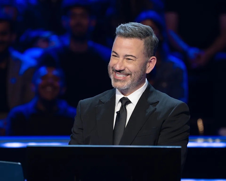 A person in a suit smiles while standing behind a podium on a stage, suggesting a television or talk show setting