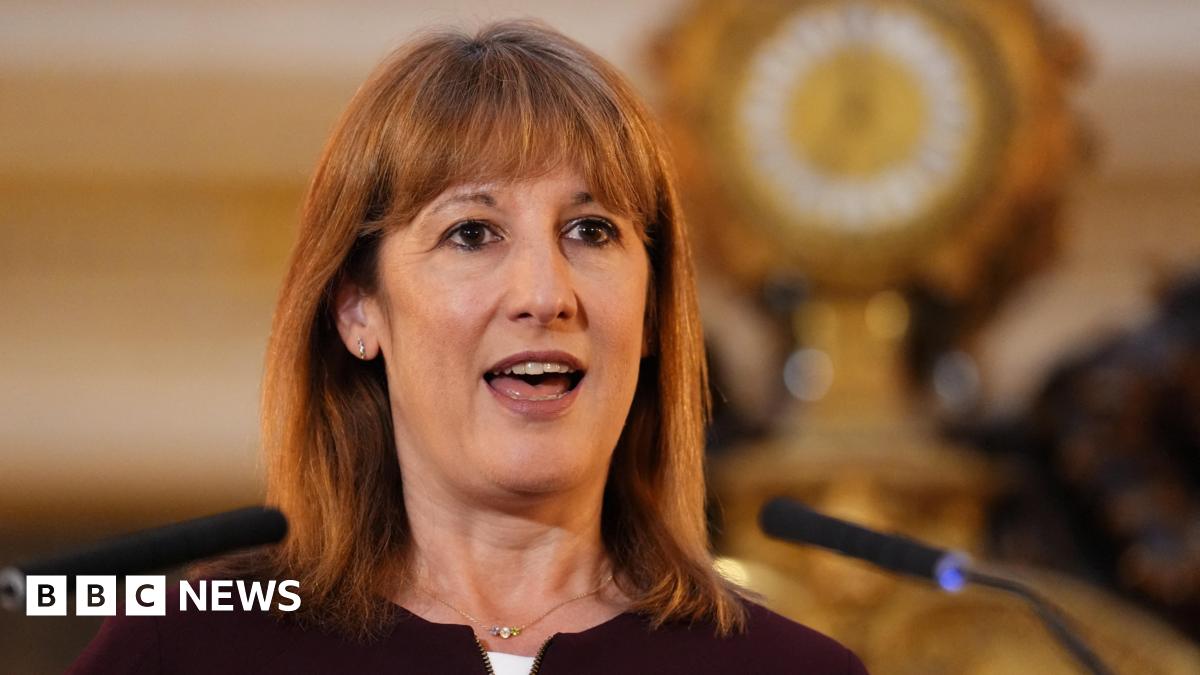 A woman with shoulder-length brown hair and a fringe speaks into a microphone, with a gold clock featuring prominently behind her. She wears a black cardigan and a gold necklace, with small silver hoop earrings.