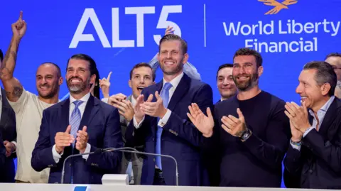Bloomberg/Getty Eric and Donald Trump in dark blue suits and light blue ties clap hands at the Nasdaq stock exchange surrounded by other executives from World Liberty Financial in August 2025