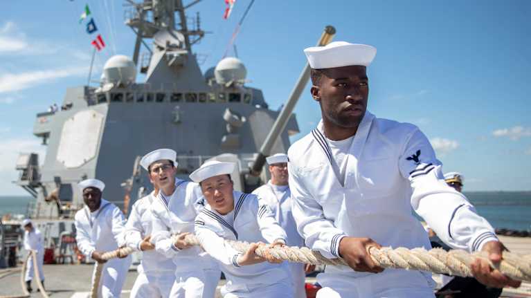 Half a dozen sailors in white uniforms pulling a rope in front of a grey naval ship.