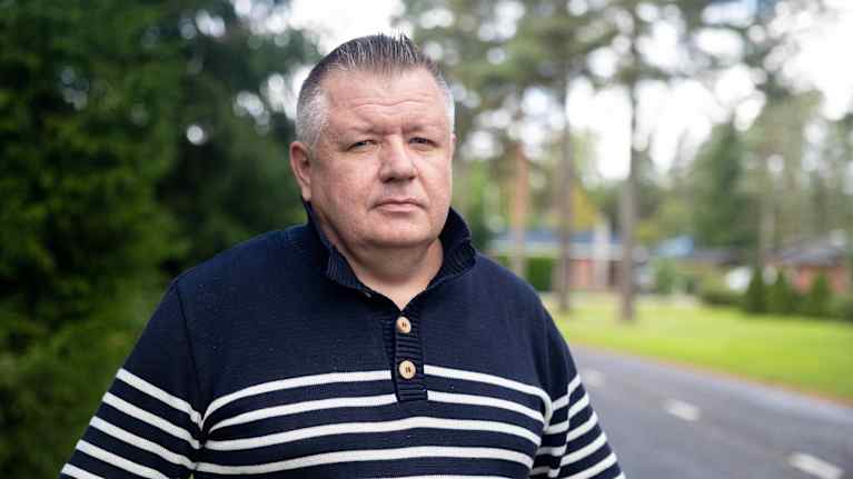 Raitapaitainen mies seisoo tien laidassa ja katsoo kameraan.