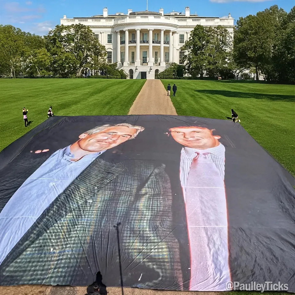 A large banner on the White House lawn displaying two images of Donald Trump in suits and ties. The White House, a white building with columns, is visible in the background with a pathway and green lawn. Several people are scattered on the lawn near the banner.