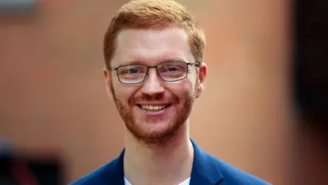 Getty Images Ross Greer, a ginger man with glasses, smiles at the camera. He is wearing a blue jacket and white t-shirt. 