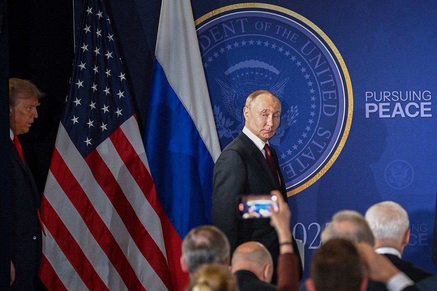 Putin walks past a seal for the President of the United States with the words "Pursuing Peace" on the backdrop behind him. The flags of Russia and the United States are to his left. Trump is seen entering at far left. Members of the media including one holding a phone to document the scene are in the foreground.