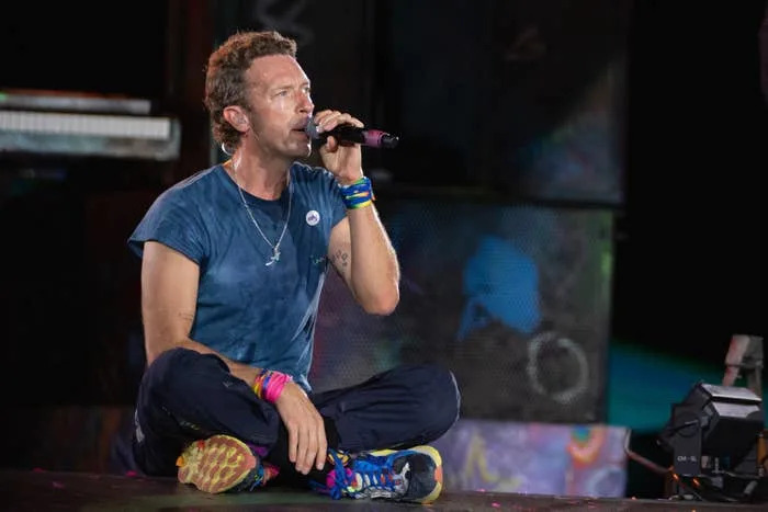 Chris Martin seated on stage, wearing a casual t-shirt, colorful shoes, and wristbands, holding a microphone while performing