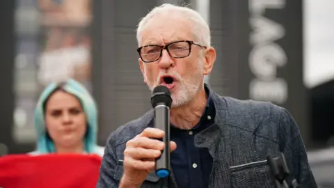 PA Media Jeremy Corbyn, a man with short grey hair, glasses and a grey beard, speaks into a microphone, held in his right hand. He is standing outside, with a woman holding a red sign and a large building in the background. He is wearing a dark jacket and shirt. 