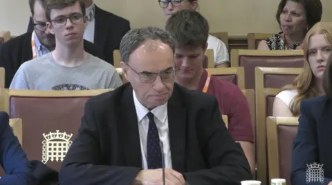 House of Commons Andrew Bailey wears a black suit jacket, white shirt and navy blue tie while sitting at a committee table. there are several people sitting in chairs in three rows behind him.
