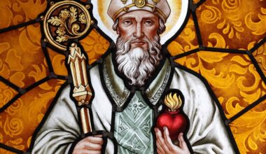 Photo of a stained glass window depicting Saint Augustine of Hippo. He is wearing a white robe and holding in his right hand a staff with some kind of flora in a circle at top. In his left hand, he is holding a heart (the shape, not anatomical) with flames coming out of the top.