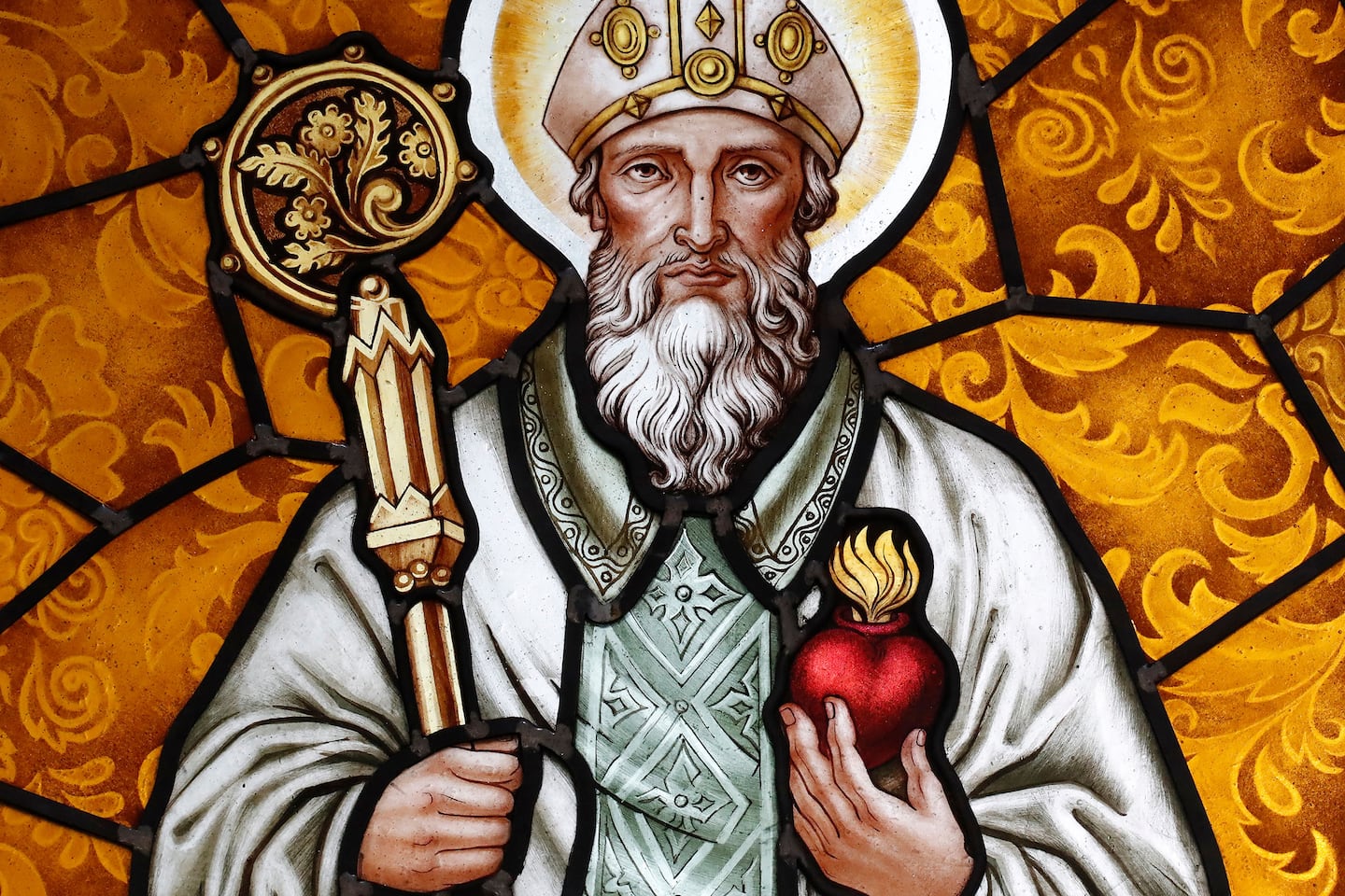 Photo of a stained glass window depicting Saint Augustine of Hippo. He is wearing a white robe and holding in his right hand a staff with some kind of flora in a circle at top. In his left hand, he is holding a heart (the shape, not anatomical) with flames coming out of the top.