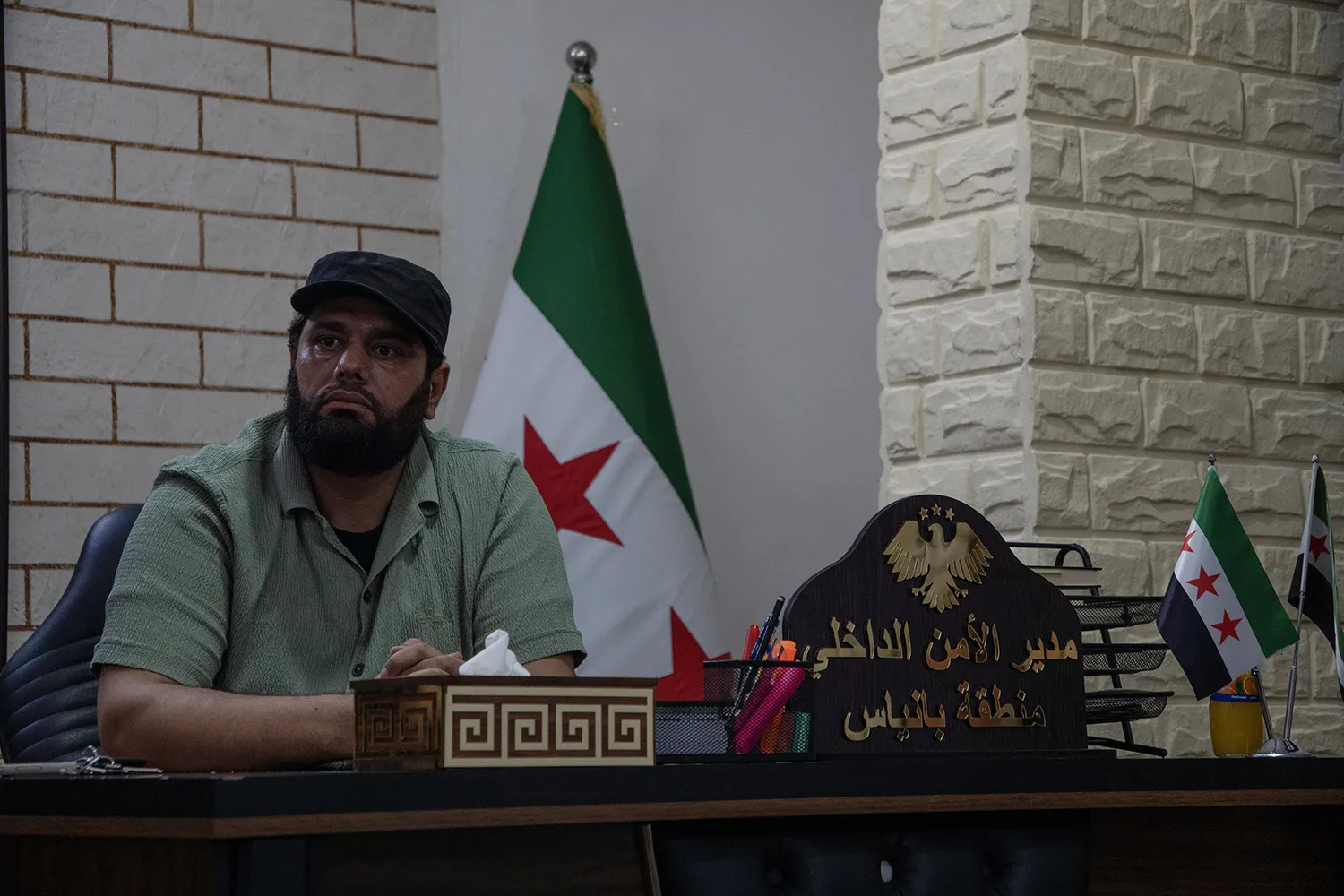 A man in a cap sits at a desk. Behind him is a flga on a stand. A plague with Arabic letters is on the desk in front of him along with office supplies and two smaller flags.