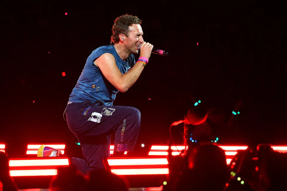 Chris Martin performs energetically on stage, wearing casual pants and a t-shirt, with colorful wristbands, amidst bright stage lights and cheering fans
